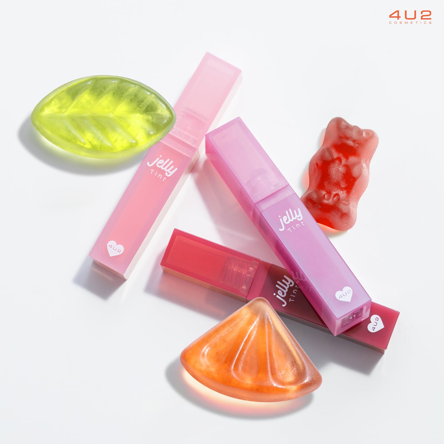 Flat-lay image of 4U2 Jelly Tint lip products in vibrant pink and purple packaging, styled alongside colorful gummy candy, including a green leaf, orange wedge, and red gummy bear, on a bright white background. The playful composition highlights the product's fun and youthful aesthetic.